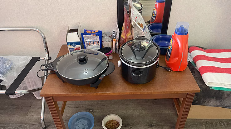 Small table in a hotel room used for storing large cooking appliances like a crockpot and electric skillet, which also doubles as a dog supply station with food and treats.